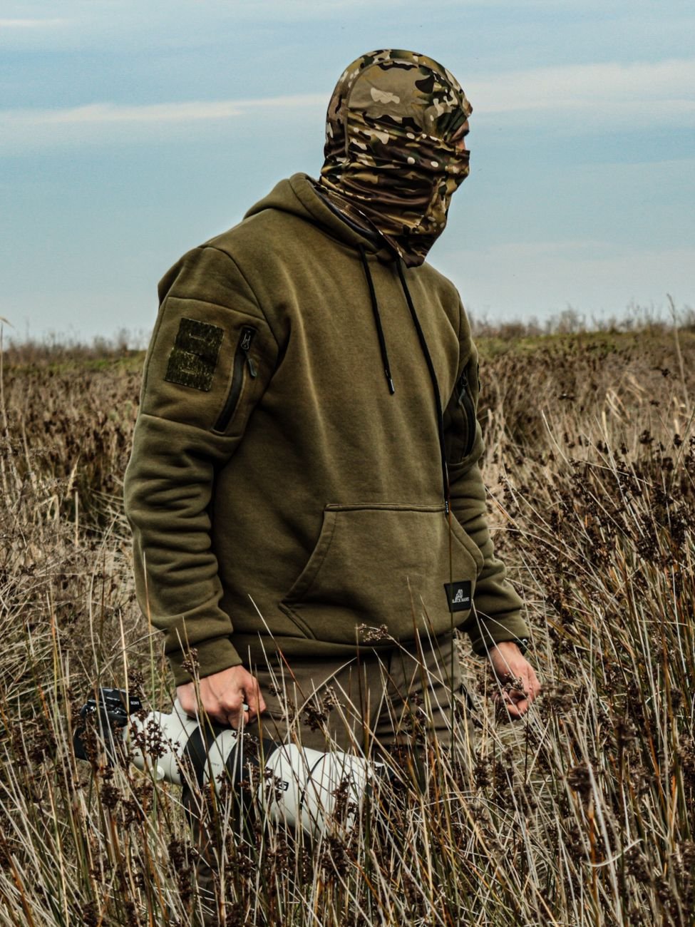 A man wearing camouflage and a hoodie stands in a grassy field